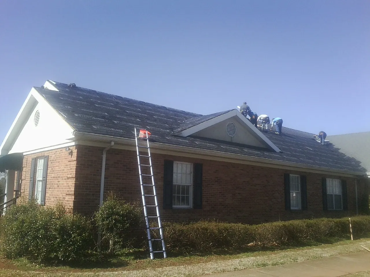 Crew tearing shingles off a brick ranch roof with a ladder set — Metal Roof Repair work by our crew in Manvel
